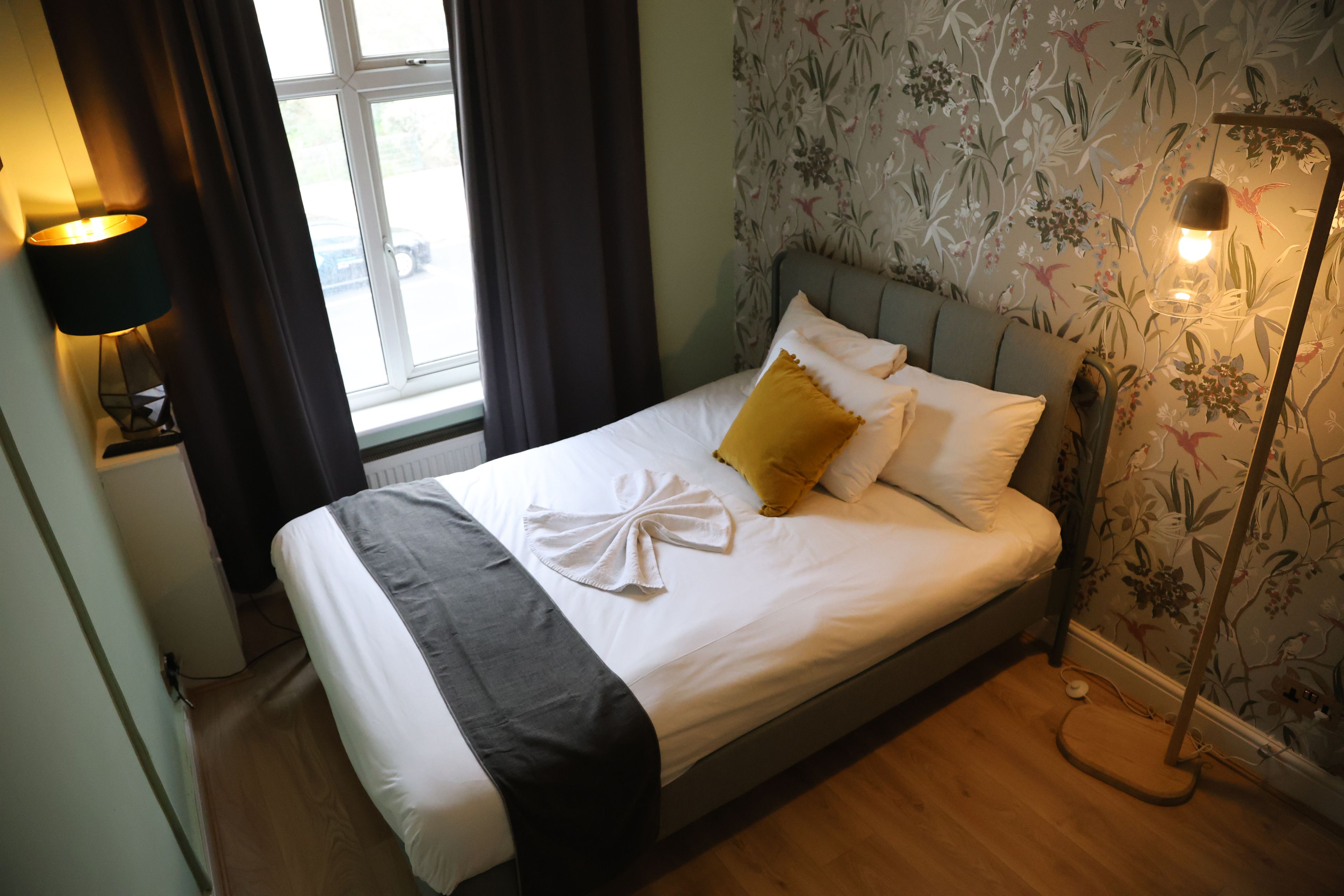 A neatly made bed with a white duvet, a grey throw blanket, and a yellow pillow. There is a bedside lamp on the left and a floor lamp on the right. The room has floral wallpaper and a window with curtains.