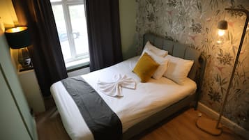A neatly made bed with a white duvet, a grey throw blanket, and a yellow pillow. There is a bedside lamp on the left and a floor lamp on the right. The room has floral wallpaper and a window with curtains.