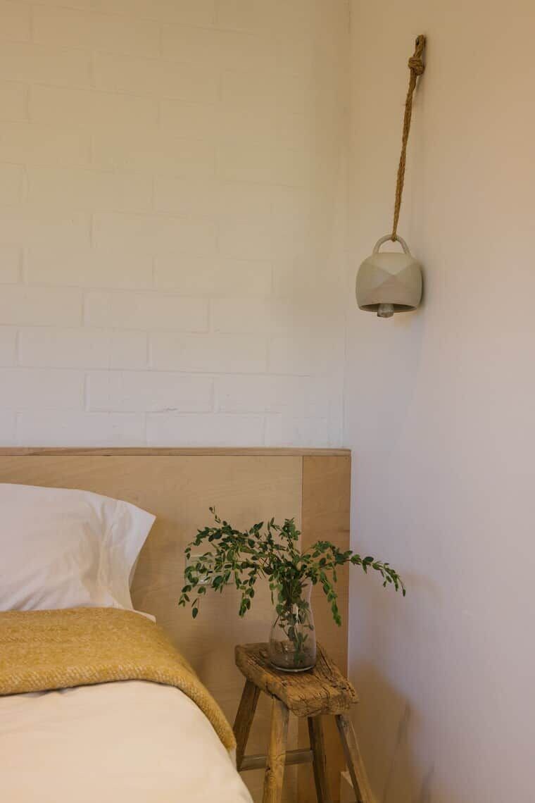 Une chambre avec une tête de lit en briques blanches, un lit recouvert de linge de lit blanc, un petit tabouret en bois sur lequel est posée une plante en pot, et une lampe fixée au mur.