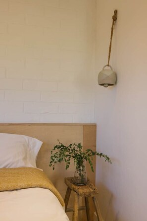 Une chambre avec une tête de lit en briques blanches, un lit recouvert de linge de lit blanc, un petit tabouret en bois sur lequel est posée une plante en pot, et une lampe fixée au mur.