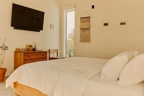 A bedroom with a bed, a TV mounted on the wall, a wooden dresser, and a macrame wall hanging. - Quail Springs Ranch | Pool & Spa (Joshua Tree)