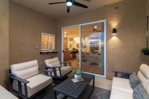 Un área de estar exterior moderna con ventilador de techo, puerta de vidrio que da a un espacio interior y una pequeña mesa con una planta.