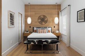 A modern bedroom with a wooden headboard, a large bed with multiple pillows, bedside tables, a bench, and wall art. - The Fairway House Scottsdale (Scottsdale)