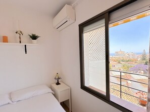 Ein Schlafzimmer mit Bett, Nachttisch und einem Fenster mit Blick auf Gebäude und das Meer.