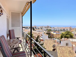 Ein Balkon mit gestreiften Kissen und einem Glastisch, mit Blick auf eine Küstenstadt mit terrakottafarbenen Dächern und blauem Himmel.