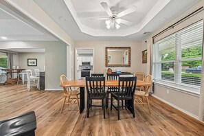 Un coin repas avec une table et des chaises en bois, un ventilateur de plafond et une vue sur l’extérieur grâce à de grandes fenêtres.