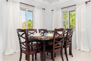 A dining area with a round table, six wooden chairs, and white curtains. - Richmond Retreat Gated Pool Beach Access (St. Ann's Bay)