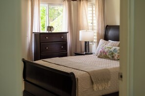 Quarto com cama, mesa de cabeceira, cômoda e janela com cortinas.