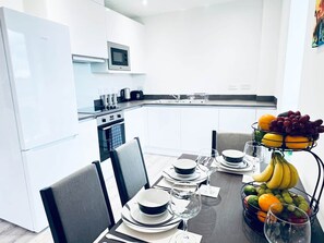 A modern kitchen with white cabinetry, a microwave, and a dining area with a table set for two, including plates, glasses, and a fruit basket. - 2BR Apt Near Birmingham City Centre | Sleeps 6 (Birmingham)