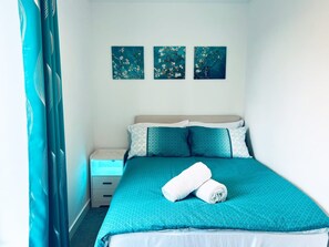A neatly made bed with a teal bedspread and pillows, a white bedside table, and three wall-mounted artworks. - 2BR Apt Near Birmingham City Centre | Sleeps 6 (Birmingham)