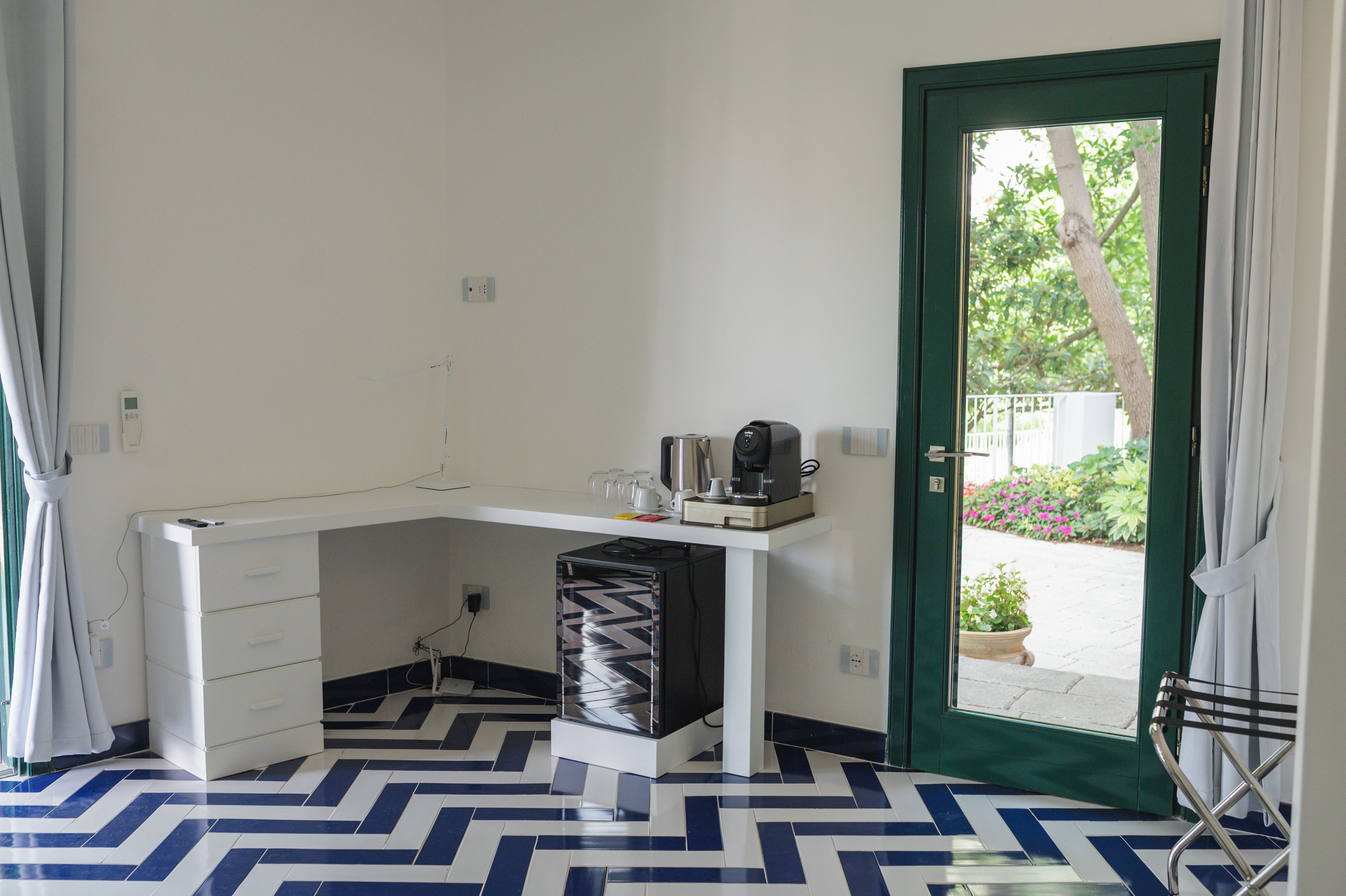Una camera compatta con una scrivania, una sedia e una macchina per il caffè. Una porta dà accesso a una zona esterna con piante.