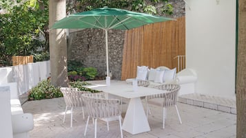 Een terras buiten met een tafel en stoelen, een groene parasol en een stenen muur op de achtergrond.