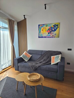 A living room with a grey sofa, two pillows, a woven basket on a wooden table, a colorful abstract painting on the wall, and a window with curtains.