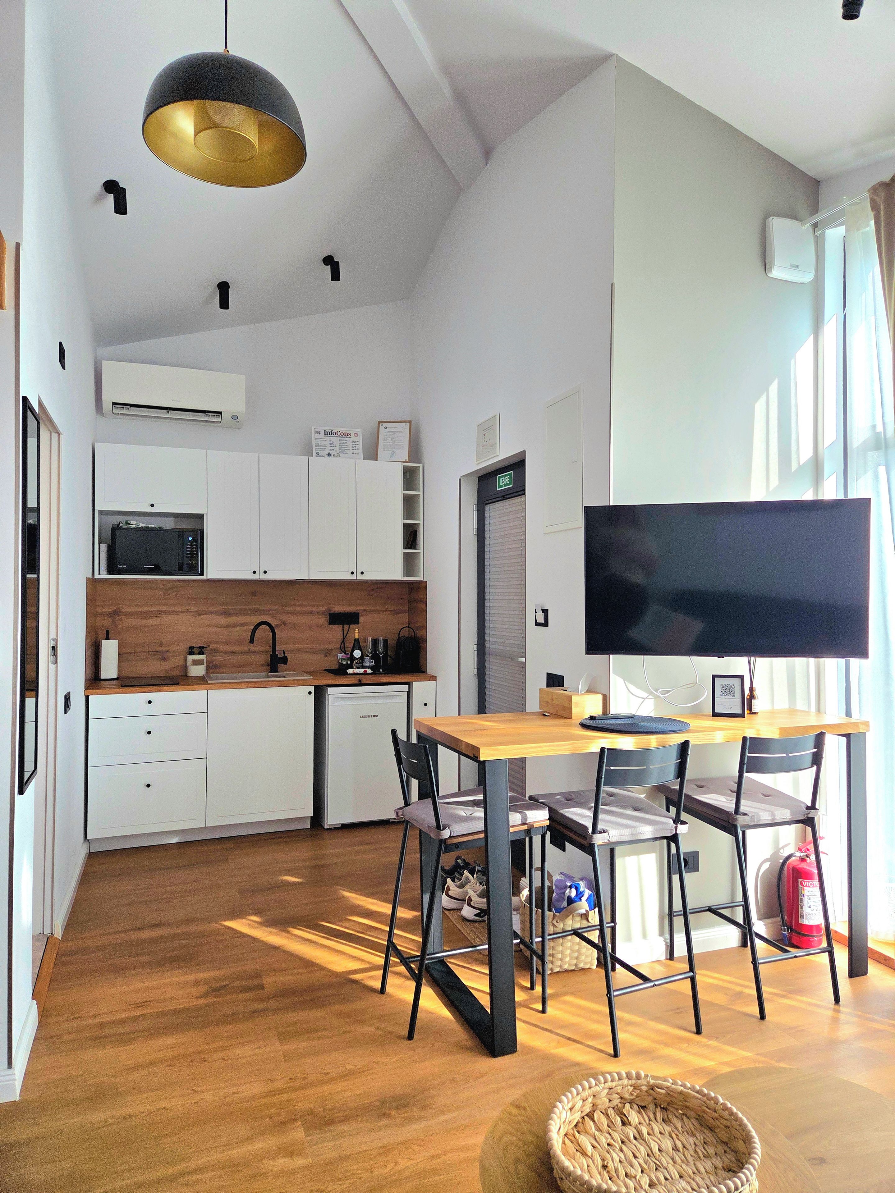 Une cuisine moderne avec des meubles blancs, un four à micro-ondes et un évier. On y trouve également un coin repas avec une table et des chaises, ainsi qu’un grand téléviseur à écran plat fixé au mur.