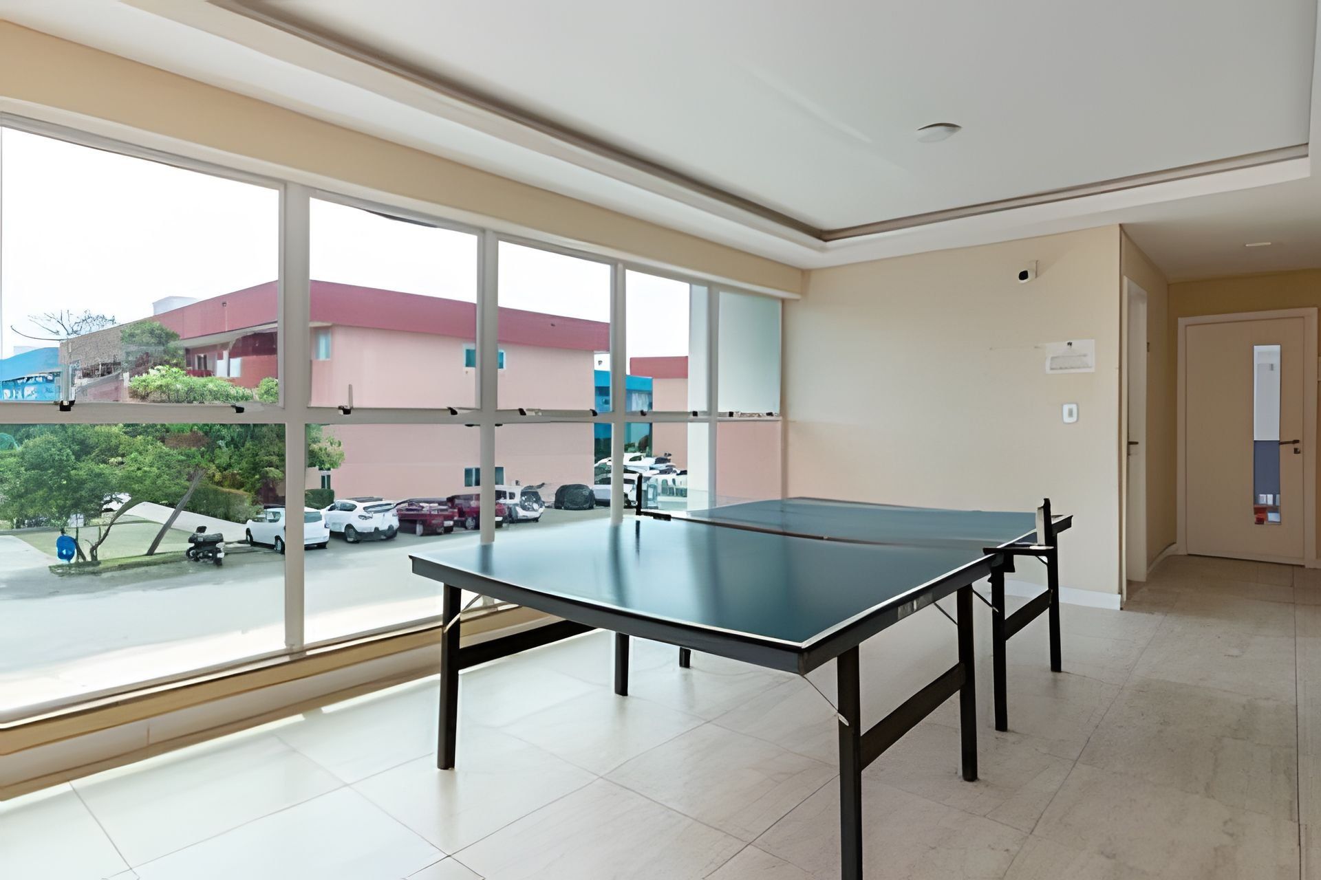 A room with a ping pong table, large windows, and a view of parked cars and buildings outside.