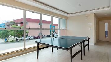 A room with a ping pong table, large windows, and a view of parked cars and buildings outside.