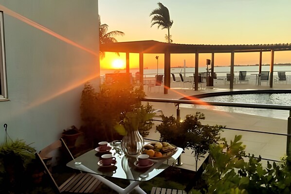 A balcony with a table set for tea, overlooking a beach and a sunset. - Apartamento Pôr do Sol (Arraial do Cabo)