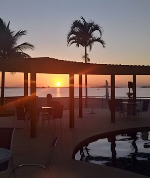 A beachside pool area with a sunset view, lounge chairs, and a covered seating area. - Apartamento Pôr do Sol (Arraial do Cabo)
