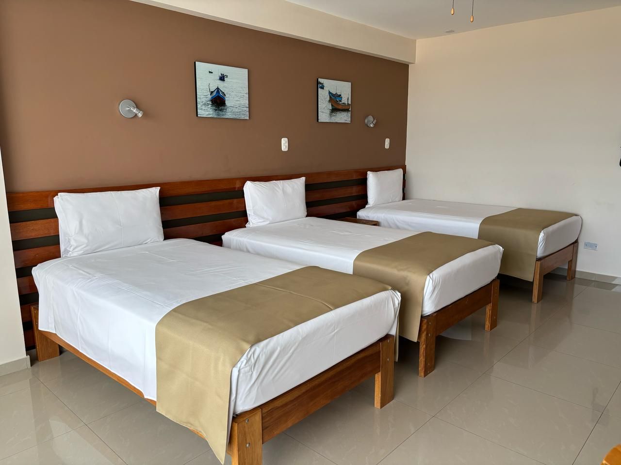 Quarto de hotel com três camas de solteiro, cabeceira de madeira, roupa de cama branca e duas luminárias fixadas na parede.
