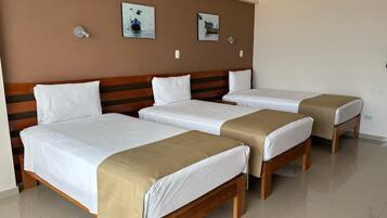 Quarto de hotel com três camas de solteiro, cabeceira de madeira, roupa de cama branca e duas luminárias fixadas na parede.