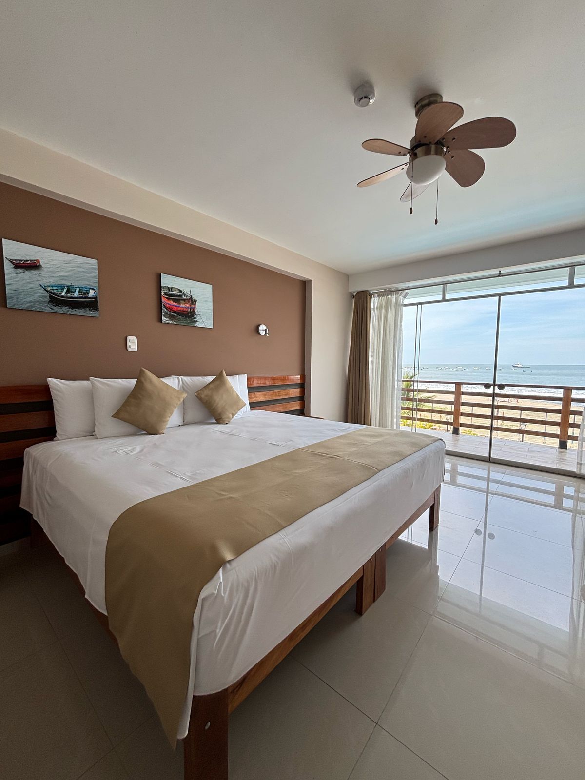 Habitación de hotel con cama, ventilador de techo, ventana grande con vista al mar y dos cuadros colgados en la pared.