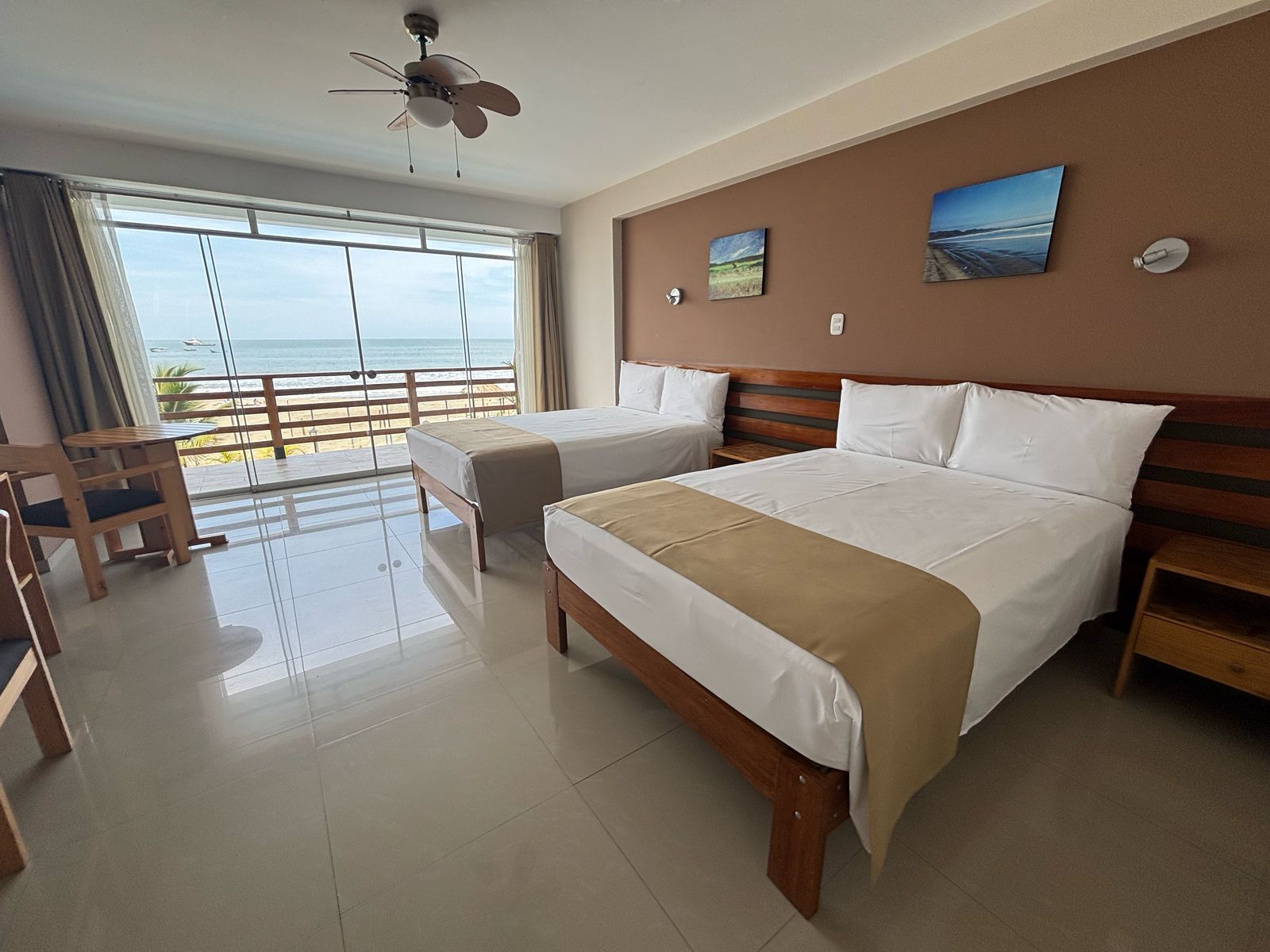 Ein Hotelzimmer mit zwei Betten, einem Deckenventilator, Blick auf den Strand und einem Holzstuhl.