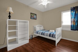 Une chambre avec un lit blanc, une étagère blanche, un ventilateur de plafond et une fenêtre avec des rideaux.
