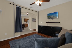 Ein modernes Wohnzimmer mit Deckenventilator, einem flachbildfernseher an der Wand, einem dunkelgrauen Sofa und einem blauen Teppich.