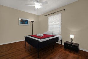 Ein Schlafzimmer mit einem Bett, einem Nachttisch, einer Stehlampe, einem Deckenventilator und einem Fenster mit Jalousien.