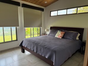 A bedroom with a large bed, a nightstand, a lamp, and a view of greenery through the windows. - Villa Aleva: Private Pool, Panoramic Views & 30+ Night Stays (Atenas)