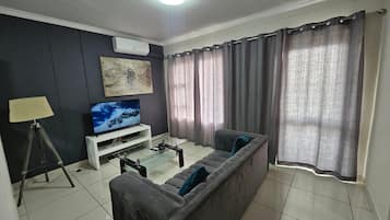 Una sala de estar con un sofá, un televisor sobre un mueble, una lámpara y cortinas.