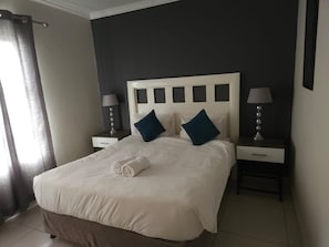 A hotel room with a bed, two bedside tables with lamps, a window with curtains, and a dark wall. - Sandton Westpoint Luxury Apartments 176 (Sandton)