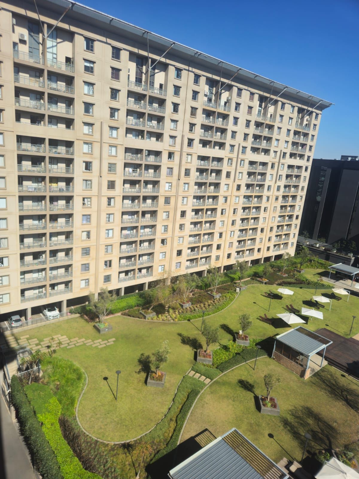 Un edificio residencial alto con múltiples balcones, un patio ajardinado con césped, árboles y mobiliario exterior.