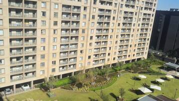 Un edificio residencial alto con múltiples balcones, un patio ajardinado con césped, árboles y mobiliario exterior.