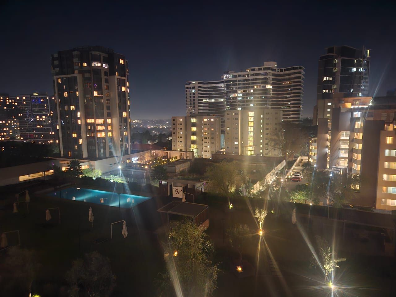 Vista nocturna de un complejo hotelero con varios edificios altos, una piscina e iluminadas calles.