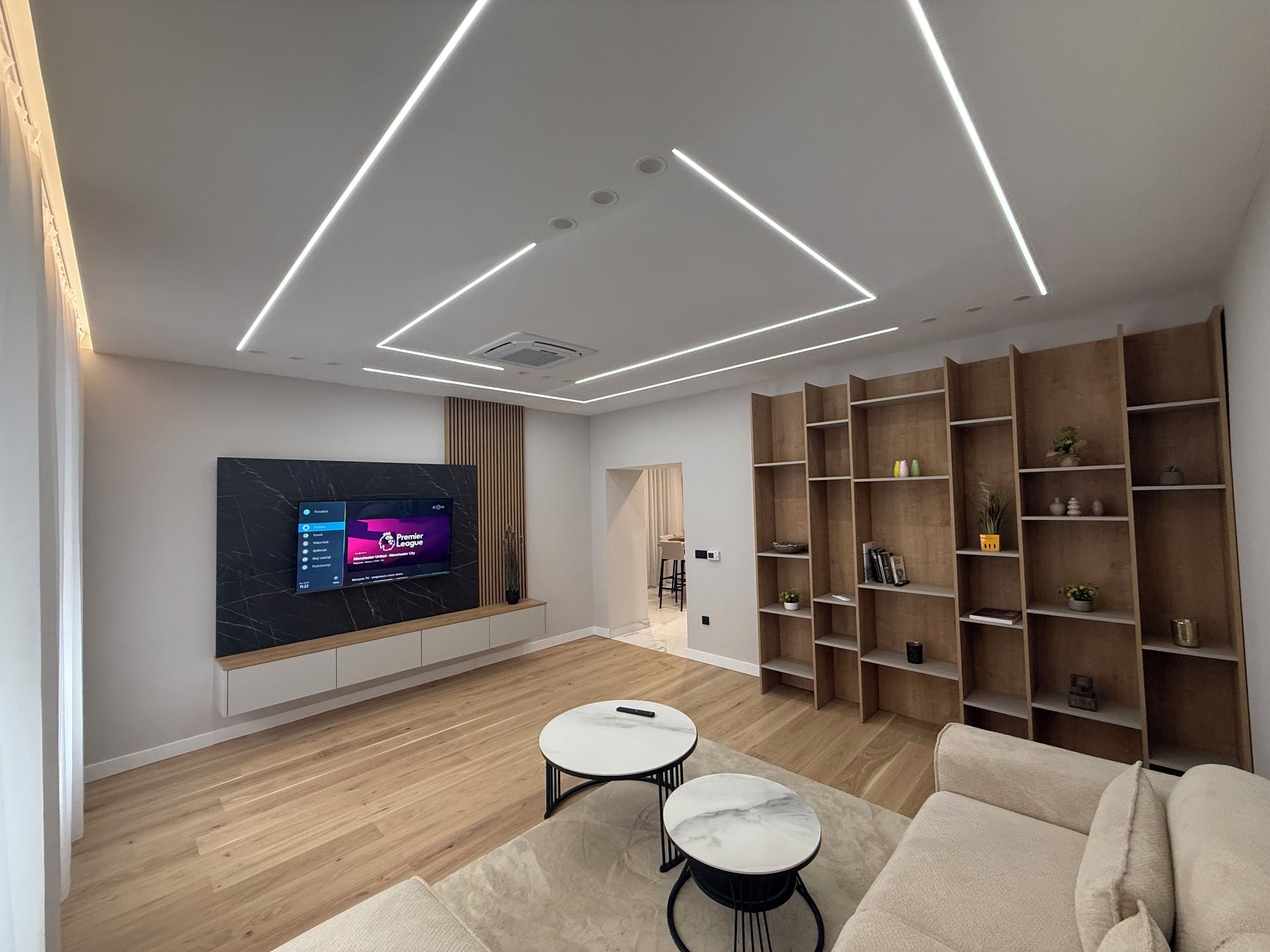 A modern living room with a flat-screen TV mounted on the wall, wooden flooring, a sofa, two round tables, and built-in wooden shelving.