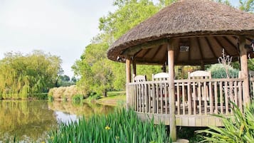 A wooden gazebo with a thatched roof, situated by a calm lake, surrounded by lush greenery.