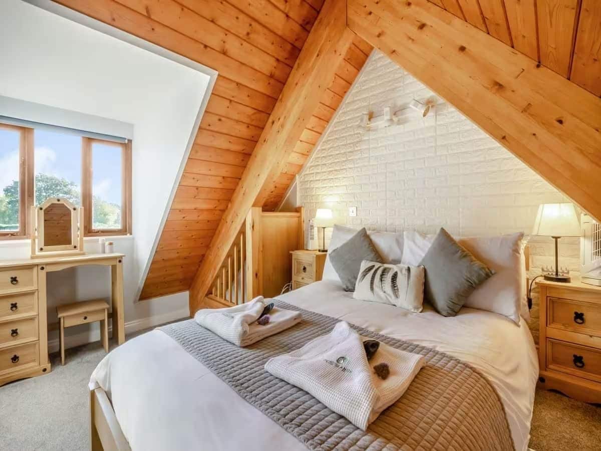 A bedroom with a sloped ceiling, a bed with pillows, a bedside table, and a desk with drawers.