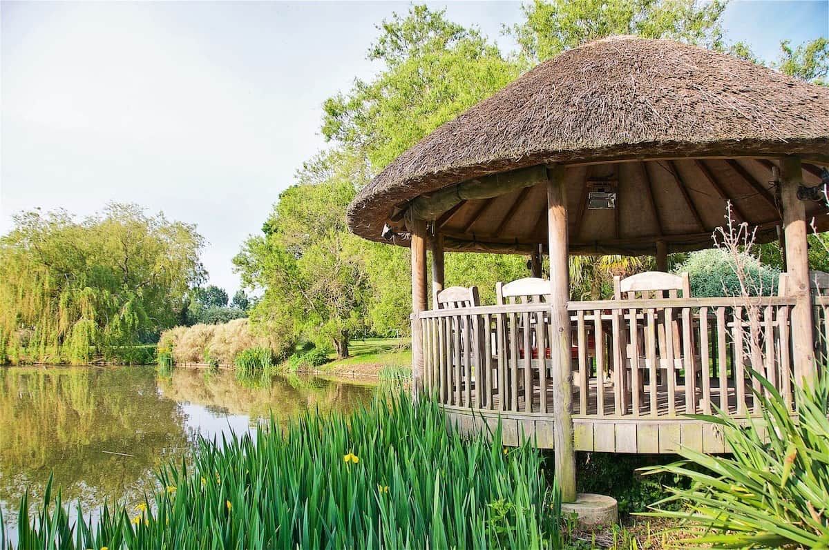 Un gazebo in legno con tetto di paglia, affacciato su un lago tranquillo, circondato da vegetazione lussureggiante.