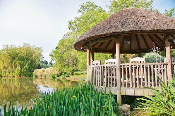 A wooden gazebo with a thatched roof, situated by a calm lake, surrounded by lush greenery.