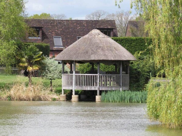A gazebo with a thatched roof, surrounded by water and greenery, with a house in the background.