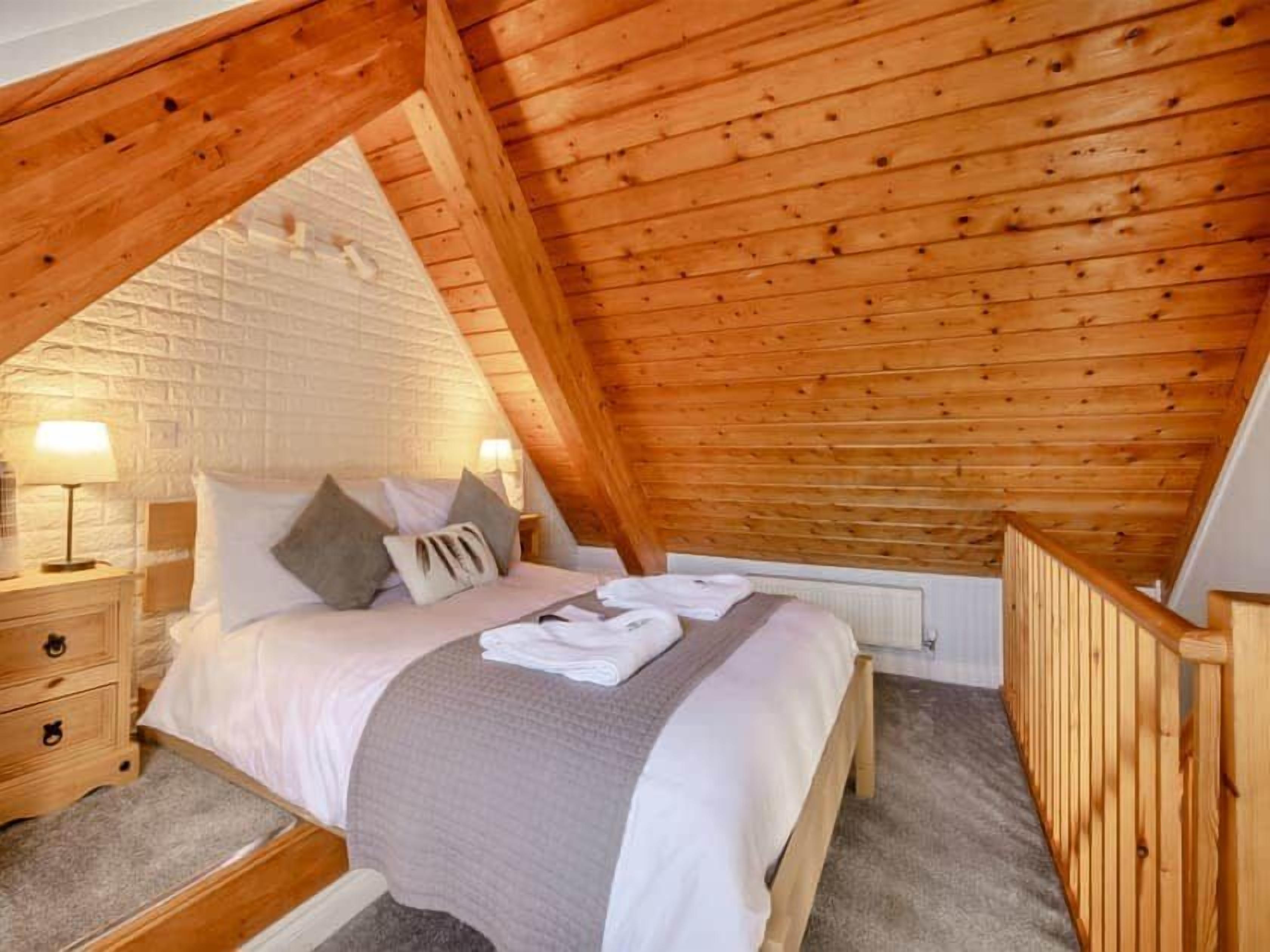A bedroom with a wooden ceiling, a bed with pillows, a nightstand, and a lamp.