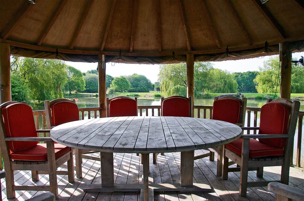 A wooden deck with a round table and red chairs, overlooking a park with trees and a body of water.