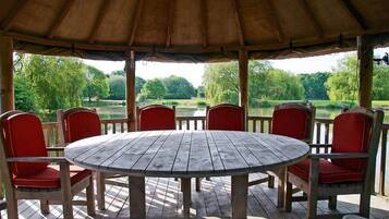 A wooden deck with a round table and red chairs, overlooking a park with trees and a body of water.