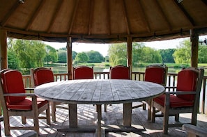 A wooden deck with a round table and red chairs, overlooking a park with trees and a body of water.