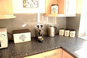 A kitchen with a bread maker, a kettle, and jars of biscuits, coffee, tea, and sugar on the counter.