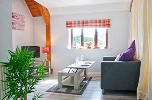 A living room with a television, a table with glasses, a sofa with a cushion, a window with curtains, and a plant.