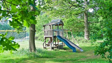 A wooden play structure with a slide in a grassy area surrounded by trees.