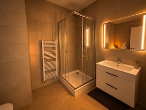 Un baño moderno con ducha envuelta en vidrio, lavamanos blanco e iluminación integrada en el espejo.