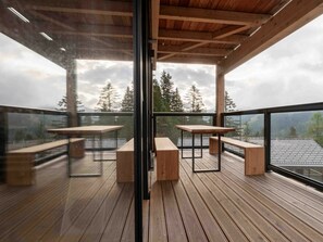 An outdoor deck with wooden tables and benches, glass railings, and a view of trees and mountains. - Cabin in Turrach With Sauna and Balcony (Stadl-Predlitz)